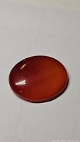 Close-up photo of a deep orange-red oval cabochon carnation gemstone against a light background showing its smooth polished surface and natural inclusions.