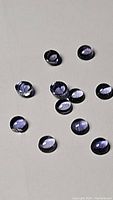 Twelve small oval-shaped purple iolite gemstones scattered on a grey surface. Stones exhibit polished, faceted cuts with light reflections.