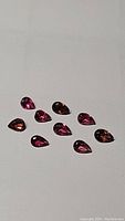 Ten pear-shaped faceted deep pink to purple tourmaline gemstones arranged on a white surface showing color and clarity.