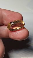 Oval cut golden citrine gemstone held between thumb and forefinger, showing facets and transparent, vivid yellow coloration.