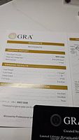 Certification papers and GRA warranty card displayed with grading report showing 1 carat, VVS1 clarity, D color, excellent cut round brilliant moissanite.