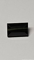 Rectangular baguette-cut green tourmaline gemstone with dark green color and polished facets, shown on flat neutral background.