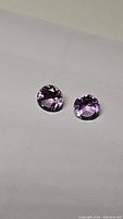 Pair of round diamond cut natural amethyst stones on white background showing size, color, and clarity.