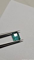 Close-up image of a natural emerald held by tweezers showing the emerald cut and rich green color on white background.