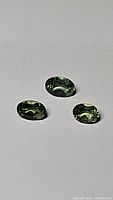 Three bright green oval faceted Apatite gemstones displayed on a flat surface, showing their size and cut.