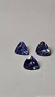 Three natural tanzanite gemstones cut in trillion shape, displayed on a neutral background, showing their size and color variation.