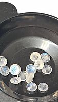 Top close-up view of multiple small calibrated round cut rainbow moonstone gemstones in a black container, showing blue flashes on translucent white stones.