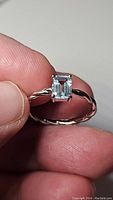 Close-up photo of a sterling silver ring with a rectangular emerald-cut natural sky blue topaz gemstone held by four prongs. The band has a twisted design and rhodium plating.