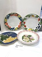 Four ceramic/porcelain tableware items: two plates with fruit borders, one floral bowl, one blue plate with corn design.
