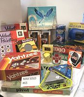 Photo of the lot showing assortment of board games (Yahtzee, UNO), velvet painting kits, jigsaw puzzles, novelty collectible tins and mugs, figurines and radio alarm clock.