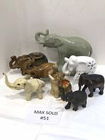 Wide shot showing all nine elephant figurines of various sizes and materials arranged on a white surface against a white wall.