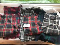 Photo showing 7 folded kilts in different tartan patterns including red, black, green, white, and navy, arranged side by side.