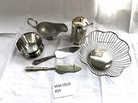 Metal gravy boat, footed bowl, jam jar cover with spoon, decorative wire bowl, and two cake servers arranged on white background