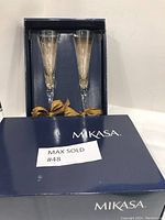 Two Mikasa crystal champagne flutes in original navy blue Mikasa box with gold ribbon bows. The box shows wear but the flutes appear like new.