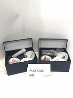 Two navy blue boxes, each containing a set of demi-tasse cups and saucers with floral designs inside, wrapped in protective packing material.