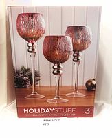 Front photo of new in box Holiday Stuff tall glow glass stem candle holder set featuring three candle holders with textured reddish-orange bowls and clear stems on box cover.