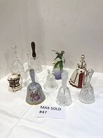 All 8 bells displayed on a white surface, showing variety in materials including clear glass, cut glass, ceramic, and a decorative bird bell.