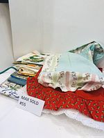 Lot of vintage linens including aprons and tea towels stacked on a flat surface with a Max Sold sign.