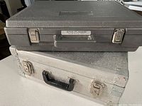 Two gear boxes stacked, upper is a black plastic Plasbox case with label, lower is a metal strong box with black handle and locking clasps