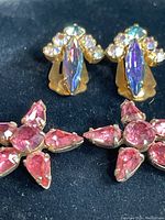 Close-up of vintage clip-on earrings with iridescent marquise and small round rhinestones alongside pink star-shaped rhinestone brooches on black background.