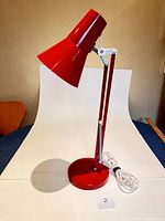 Red articulating desk lamp showing side view with white backdrop, adjustable arm extended upward, round base, and power cord wrapped on the side.