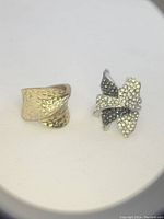 Two rings displayed side-by-side on a white surface, showing the hammered gold-tone band and the rhinestone floral ring.