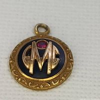 Front side of circular pendant with gold filled script initials GMC and a small red ruby stone in center with ornate border detail