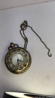 Front view of the brass-toned quartz pocket watch with visible clock face and attached chain.