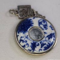 Porcelain pendant showing one side with blue floral design, metallic center, and silver frame; visible cracks and chips.