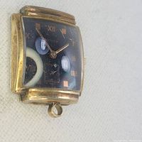 Close-up of gold-toned rectangular pendant watch with black dial and Roman numeral markers, showing the face and small seconds sub-dial.