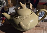 Close-up angle of one light green ceramic teapot showing sculpted hen-shaped lid handle and embossed chicken on pot body with vine design decoration.