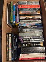 Top-down view inside a box showing assorted DVD sets with visible titles Lost, Dilbert, The Simpsons, Clerks, Heroes, Sherlock, Game of Thrones.