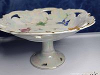Side angle view of the iridescent serving pedestal showing scalloped openwork edge with painted golden and colorful leaf and butterfly motifs.