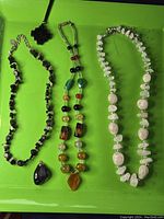 Photo showing four necklaces and one pendant on a green background, including black bead necklace, multicolored bead necklace, white/pink stone chip necklace, dark stone bead necklace, and dark teardrop pendant.