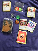 Top view showing a stack of Pokemon energy cards including fire energy, trainer cards, a sealed pack of black card protectors with an orange Pokemon design, three colorful coins in orange and green, and two Pokemon pins.
