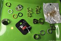 Many assorted rings on green surface including silver, gold, black, and colorful types.