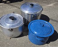 Three large lidded pots on outdoor surface, two aluminum and one blue speckled enamel