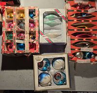 Top-down view of three vintage cardboard boxes filled with assorted glass Christmas ornaments in multiple colors including blue, gold, red, green, and pink, plus a boxed set of green teardrop shaped ornaments.