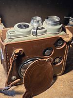 Photo showing the vintage movie camera with silver and grey metal body partially inside a brown leather case with attached lens cap.