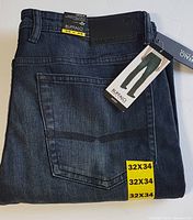 Folded pair of Buffalo David Bitton Jack-X slim straight stretch denim jeans in dark indigo blue with attached tags and labels showing size 32x34 and branding details.