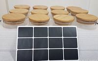 Twelve clear glass jars arranged in rows with bamboo lids and a sheet of black adhesive labels placed in front. Photo shows jar size and design clearly.