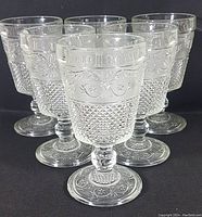Front view of all six vintage American Sandwich glass water goblets showing intricate pressed glass pattern and overall shape.