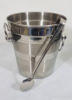 Front and side view of the stainless steel ice bucket with one handle visible and ice tongs resting against the bucket.