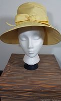 Front view of vintage yellow ladies hat with bow, displayed on a white mannequin head placed on a Simpsons branded box.