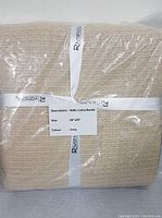 Folded new waffle cotton blanket in ivory color wrapped in plastic with label and ribbons showing brand and details.