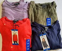 Four folded Buffalo David Bitton ladies long sleeve knit tops in lavender, olive green, red with heart pattern, and navy blue stacked on a white sofa.