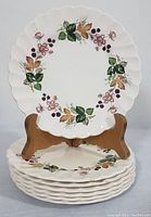 Seven side plates with floral and leaf design stacked, one displayed upright on a wood plate holder, showing pattern and edge detail.