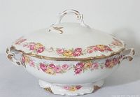 Front side view of the white porcelain serving bowl with lid featuring yellow and pink rose garlands and gilded handles.