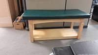 Side view of Com*Fort massage therapy table with green vinyl padded top, wooden frame, and lower shelf.