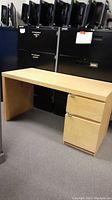 Light wood office desk with two drawers on right side, shown against background of black filing cabinets and monitors.